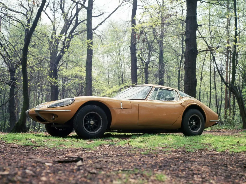 A yellow 1960s Marcos GT photographed in profile in a wooded clearing